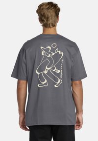Grå kortærmet t-shirt med et stort beige grafik af en person, der holder et surfbræt, med afrundede kanter og en afslappet pasform.