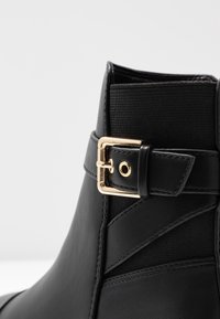 Schwarze Knöchelstiefel aus glattem Leder mit einem strukturierten elastischen Einsatz. Verfügt über einen goldfarbenen Metallschnallenakzent an der Seite.