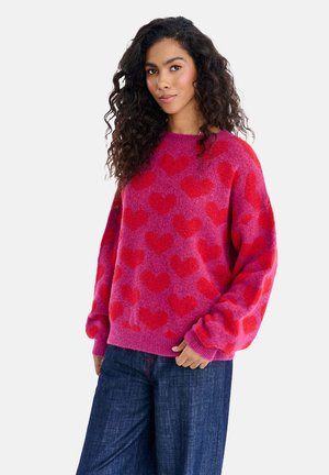 Kobieta z kręconymi włosami w różowym sweterze w czerwone serca oraz ciemnoniebieskich dżinsach, pozująca na jednolitym białym tle.