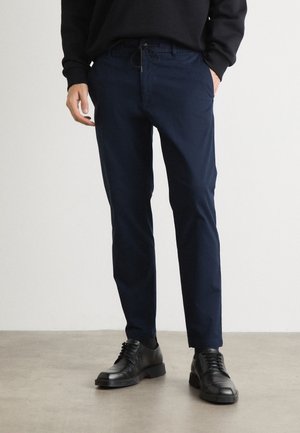 Mann trägt marineblaue, schmal zulaufende Hose mit Kordelzug in der Taille und schwarze Lederschuhe, steht auf grauem Boden.