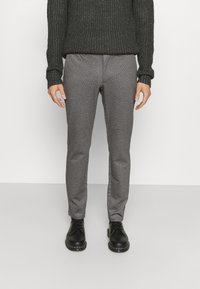 Pantalons gris à motifs en tissu texturé, assortis à un pull en tricot noir. Chaussures en cuir noir avec un bout rond.