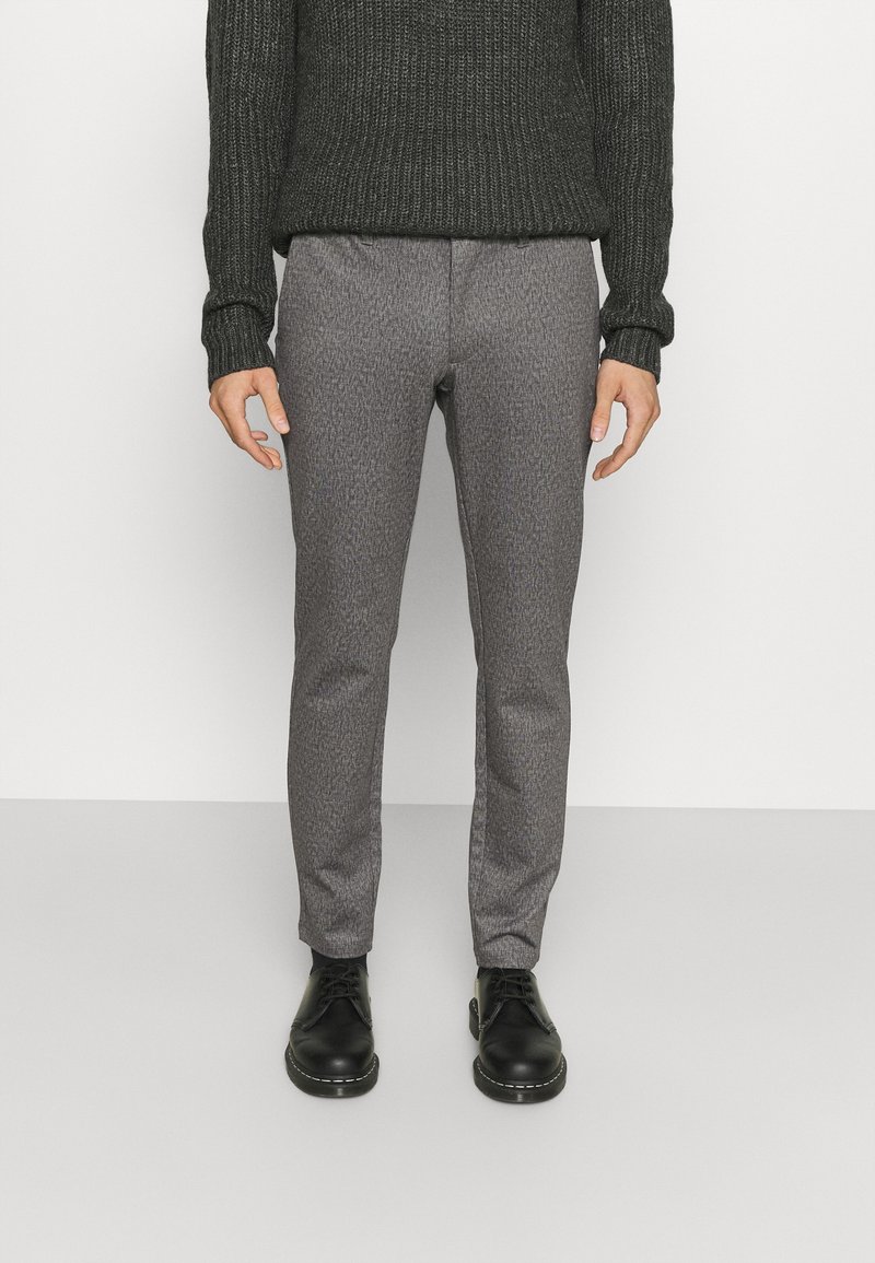 Pantalons gris à motifs en tissu texturé, assortis à un pull en tricot noir. Chaussures en cuir noir avec un bout rond.