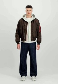 Ung mann som står, iført en mørkebrun bomberjakke over en hvit hettegenser, mørkeblå vide jeans og hvite sneakers.
