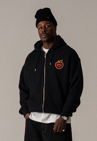 Felpa nera con cappuccio con zip frontale e tasca anteriore, con logo a mela rossa ricamato. Indossata sopra una camicia bianca, abbinata a un berretto nero.