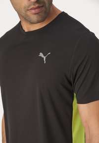 Czarny sportowy t-shirt z krótkim rękawem, wykonany z teksturowanej tkaniny, z zielonymi panelami bocznymi i białym logo Pumy na piersi.