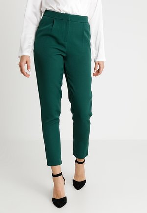 Mujer que lleva pantalones oscuros verdes de talle alto y corte ajustado, blusa blanca de manga larga y tacones negros con correa en el tobillo caminando hacia adelante.