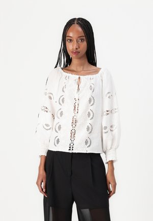 Blusa bianca con dettagli in pizzo intricati, scollo rotondo e maniche lunghe a sbuffo. Indossata sopra dei pantaloni corti neri a vita alta.