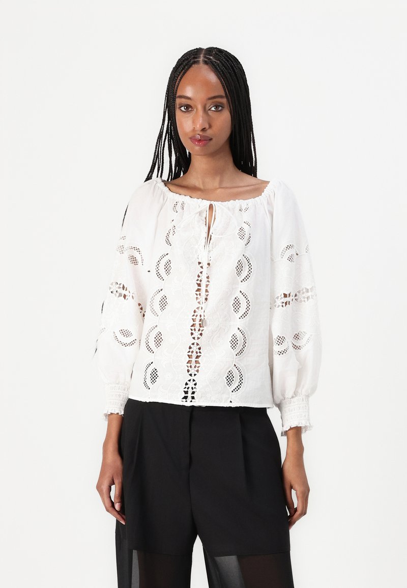 Weißes Blusenshirt mit filigranen Spitzenverzierungen, runder Ausschnitt und langen Puffärmeln. Getragen über schwarze High-Waist-Shorts.