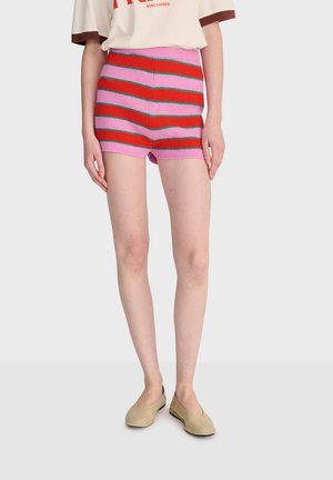 Modelka má na sebe ružové a červené pruhované pletené šortky, béžové nazúvacie topánky a krémové tričko s červeným textom na jednoduchom pozadí.