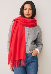Écharpe en laine rouge avec franges, portée sur un cardigan gris en maille. Le cardigan dispose d'une fermeture par boutons et d'un ourlet côtelé. Associé à un jean bleu.