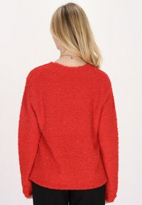 Roter strukturierter Pullover mit lockerer Passform, langen Ärmeln und rundem Halsausschnitt. Der Stoff wirkt fusselig und weich, mit einer lässigen Silhouette.