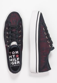 Schwarze und rote Stoffturnschuhe mit weißen Gummisohlen. Zu den Merkmalen gehören Metallösen, schwarze Schnürsenkel und ein Logo auf der Innensohle.