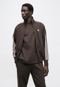 Καφέ αθλητική ζακέτα με φερμουάρ, με τρεις λευκές ρίγες σε κάθε μανίκι και ένα μικρό λευκό λογότυπο στο στήθος, συνδυασμένη με ένα αντίστοιχο πουκάμισο.