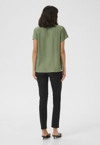 Chemise à manches courtes vert olive, coupe ample, tissu texturé ; associée à un jean noir slim et des mules noires à talons avec des nœuds.