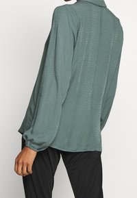 Blouse verte texturée à manches longues et col avec un subtil motif reptile, coupe décontractée. Associée à un bas noir.