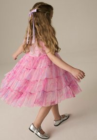 Rosa Schichtkleid mit rüschigen Stufen und Glitzer, kombiniert mit glänzenden silbernen Schuhen. Hat ein lilafarbenes Band in welligem Haar.