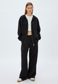 Sudadera negra con cremallera y cordones, combinada con pantalones de chándal negros holgados. Top corto blanco liso debajo. Tejido de algodón casual.