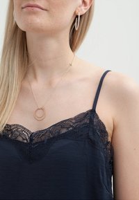 Kvinna som bär en svart spetstrimmad linne, guld hoop-örhängen och ett delikat guldhänge med två sammanflätade ringar.