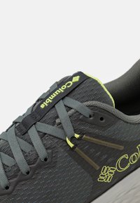 Tumši pelēki sporta apavi ar sietveida augšdaļu, zaļiem akcentiem, elastīgām auklām, pastiprinātu pirkstu daļu un teksturētu gumijas zoli.