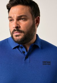 Mies, jolla on tumma hiukset ja parta, pukeutuu siniseen kauluspaitaan, jossa on rinnan kohdalla teksti "VINTAGE DENIM GEAR", katsoo hieman sivulle.