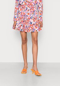 Robe florale éclatante avec des motifs roses, orange et bleus ; ourlet volanté et manches longues, associée à des sandales à talons orange sur un sol gris.