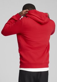 Rød sweater med hætte i blødt materiale, justerbar hætte og ribbede manchetter. Iført over en hvid skjorte, mod en ensfarvet grå baggrund.