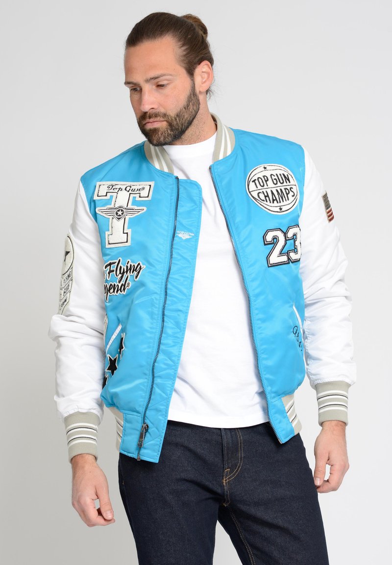 Lichtblauwe bomberjack met witte mouwen, voorzien van verschillende patches en borduursels. Belangrijke details zijn de gestreepte geribbelde kraag en manchetten.