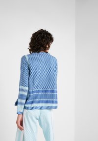 Maglietta a maniche lunghe blu con motivi vari, tra cui forme geometriche e righe; abbinata a pantaloni chiari a fantasia blu.