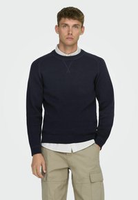 Suéter de punto azul marino con cuello redondo y puños acanalados, superpuesto a una camisa a rayas blanca y azul marino, combinado con pantalones cargo beige.