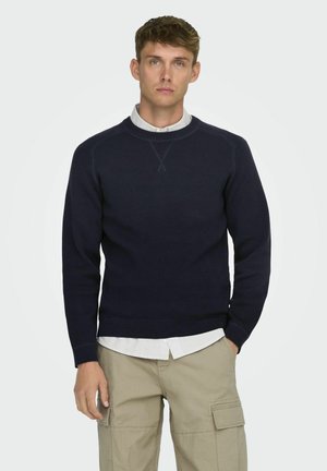 Suéter de punto azul marino con cuello redondo y puños acanalados, superpuesto a una camisa a rayas blanca y azul marino, combinado con pantalones cargo beige.