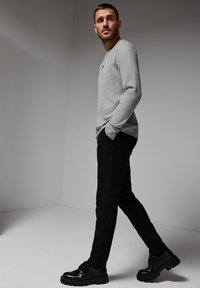 Grå långärmad tröja med rundad hals, svarta byxor och svarta skor med tjock sula. Modellen står i en neutraltonad miljö.