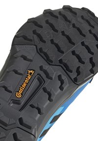 Semelle de chaussure en caoutchouc noir avec des rainures profondes, surface texturée ; présente un logo "Continental" orange et une tige en tissu bleu le long du bord.