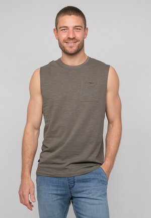 Homme souriant aux cheveux courts portant un débardeur gris avec poche poitrine et un jean bleu, une main dans la poche.