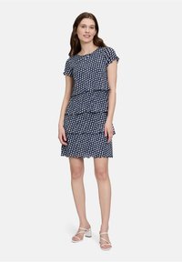 Navy-Kleid mit weißem Wellenmuster, kurzen Ärmeln, schichtweiser Gestaltung und einem Saum über dem Knie. Kombiniert mit weißen Blockabsatzsandalen.