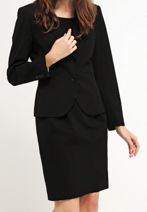 Woman wearing a fitted black business suit with a blazer and knee-length pencil skirt, standing against a plain light background.