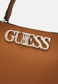 Sac à main en cuir texturé marron avec un emblème en argent "GUESS" bien en évidence en tant qu'accent matériel à l'avant.