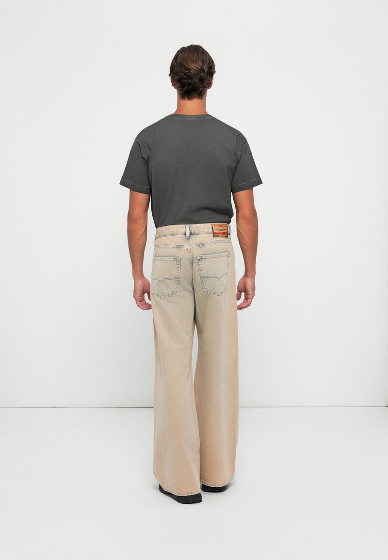 Homme vu de dos portant un t-shirt gris foncé et un jean beige large avec des poches arrière et une pièce en cuir à la taille.