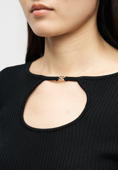 Κοντινό πλάνο στον λαιμό και το πάνω μέρος του στήθους μιας γυναίκας που φοράει μαύρο ριμπ τοπ με μικρό χρυσό κούμπωμα που συνδέει το μπροστινό ντεκολτέ σε σχήμα κλειδαρότρυπας.