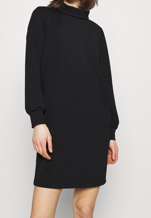 Mujer con un vestido negro mini de manga larga y cuello alto con puños, sobre un fondo liso y claro.