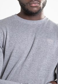 Jasnoszary dzianinowy sweter z okrągłym dekoltem i prążkowanym wykończeniem. Z przodu po lewej stronie małe, haftowane logo. Miękka tekstura.