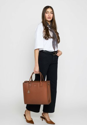 Woman wearing white top and black pants holds large brown leather tote bag, accessorized with patterned neck scarf and brown heels.