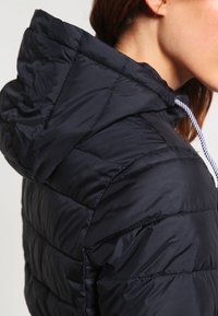 Femme portant une veste matelassée noire avec capuche et cordon blanc, vue de l'épaule vers le haut, de profil.