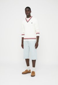 Baltas megztinis su nervūriniu raštu, raudonomis ir juodomis juostelėmis ant apykaklės ir rankogalių, derinamas su šviesiai mėlynais džinsiniais šortais ir rudais batais.