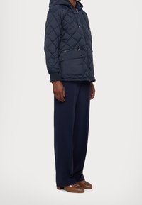 Quiltet marineblå jakke med hætte, der har diagonale syninger, to forlommer og elastiske manchetter. Bæres med vidde bukser og brune sko.