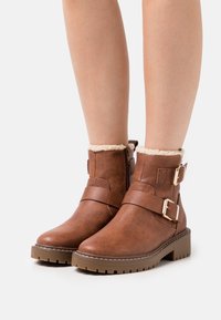 Bottines en cuir marron avec une finition texturée, dotées de deux accents de boucles et d'une douce doublure en fourrure au niveau du col. Semelle en caoutchouc épaisse.