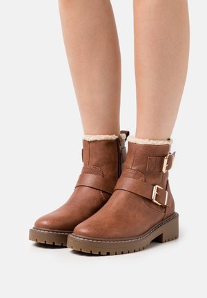 Botas de tobillo de cuero marrón con un acabado texturizado, que presentan dos detalles de hebilla y un suave forro de piel en el cuello. Suela de goma gruesa.
