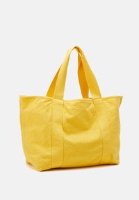 Jasnożółta torba materiałowa z szerokimi uchwytami, stojąca w pionie na tle jednolitego białego tła.