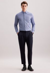 Man staat met zijn handen in zijn zakken, gekleed in een lichtblauw geblokt overhemd, zwarte broek, zwarte sokken en zwarte nette schoenen.