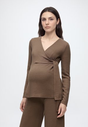 Femme enceinte aux longs cheveux foncés portant un top cache-cœur en maille marron et un pantalon assorti sur un fond clair uni.