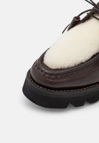 Gros plan sur une chaussure en cuir marron foncé avec une semelle noire épaisse et un panneau en polaire blanche sur le dessus près des lacets.
